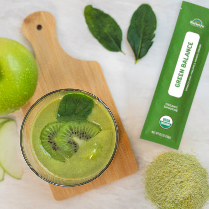 Lifestyle photo of a glass of Natierra Green Balance Organic Smoothie on a marble counter top with a smoothie stick pack, apple, and spinach on the side