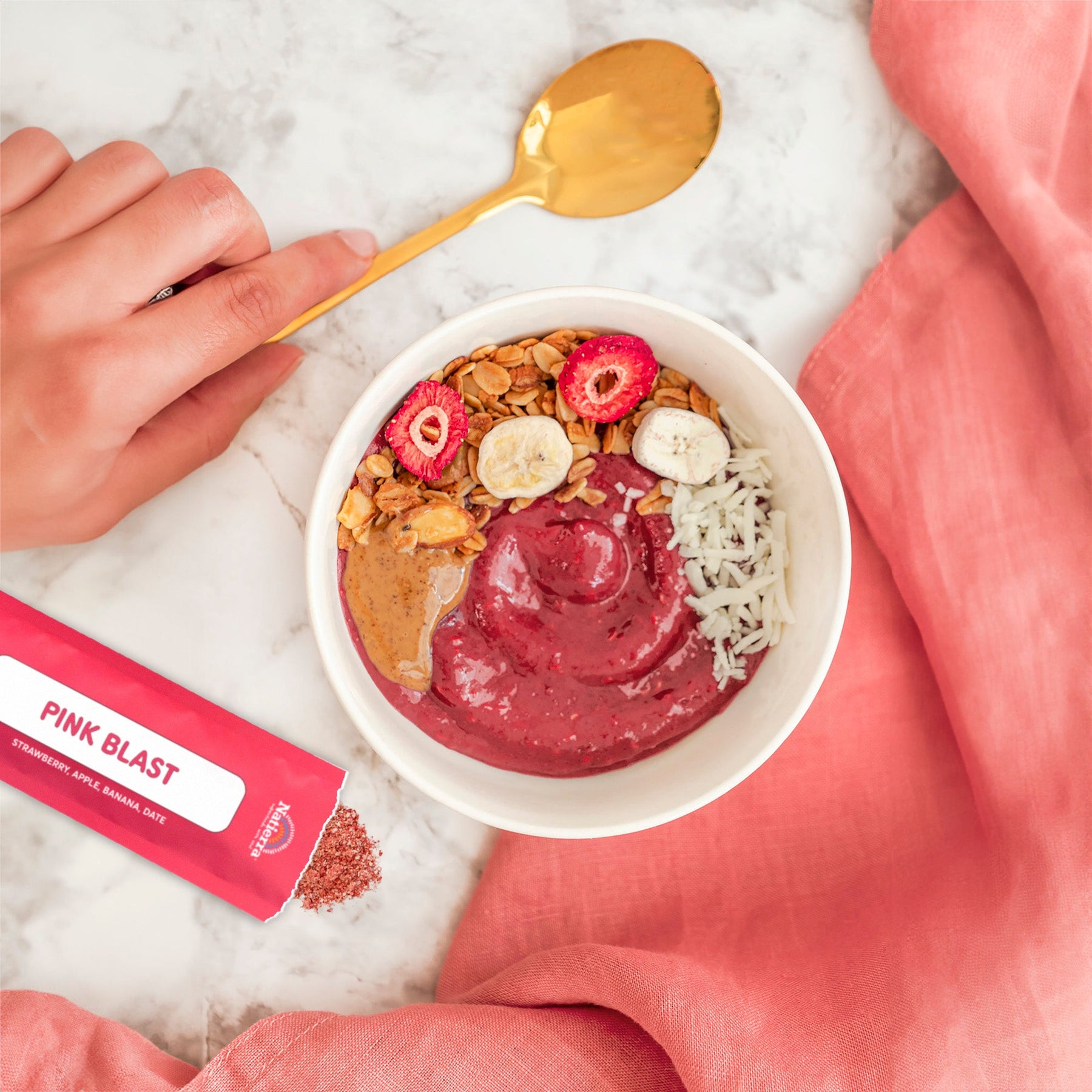 Lifestyle photo of a glass of Natierra Pink Blast Organic Smoothie on a marble counter top with a smoothie stick pack, a pink napkin and a hand holding a golden spoon on the side