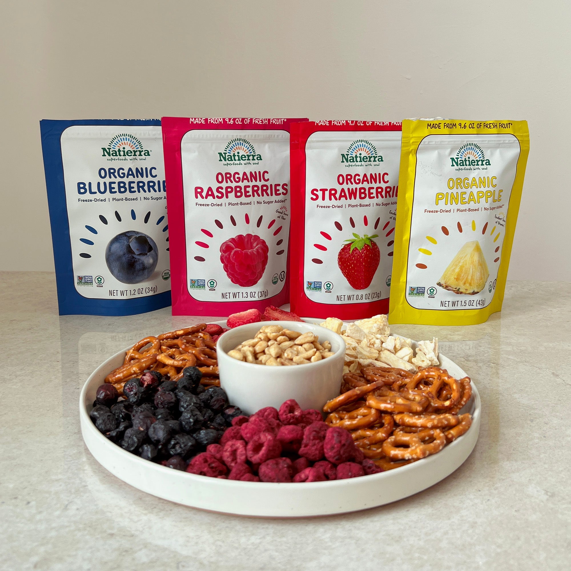Organic freeze-dried fruit bags of blueberries, raspberries, strawberries and pineapples with a plate of freeze-dried snacks, peanuts and pretzels
