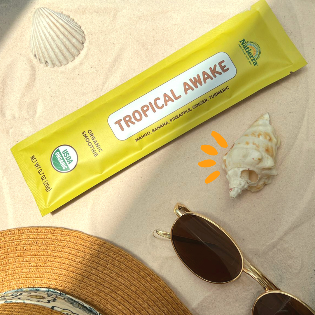 Tropical Awake smoothie blend packet on sand with shells, glasses and a sun hat next to it